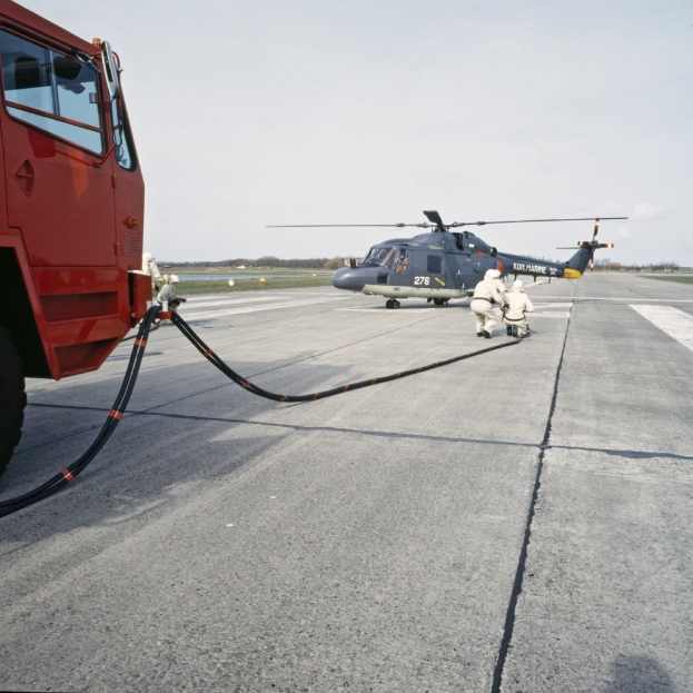 Rote Feuerwehrauto neben einem Hubschrauber auf einer Landebahn geparkt, zwei Personen in der Nähe, Bäume und ein klarer blauer Himmel im Hintergrund.