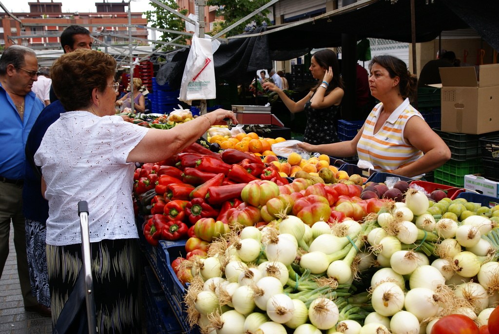 Gemüse in Plastikbehältern, eine Gruppe von Menschen, die stehen, Gebäude, Bäume, eine Kartonschachtel und der Himmel.