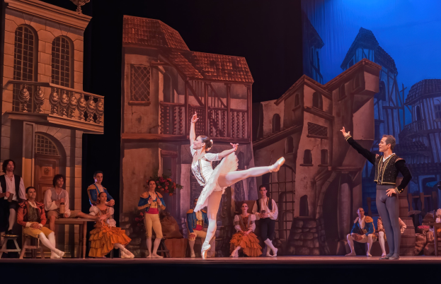 Zwei Tänzer performen auf einer Bühne im Royal Opera House in London, mit einem Publikum im Vordergrund, das auf Hockern sitzt und steht, und einem Bildschirm im Hintergrund, der Gebäudeabbildungen anzeigt.