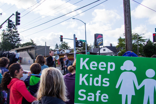 Gruppe von Menschen an einer Straße mit einem "Kinder sicher"-Schild, mit einem Mann mit Megafon im Vordergrund und Gebäuden, Bäumen und Verkehrselementen im Hintergrund.