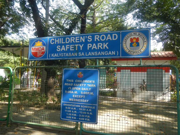 Ein von einem Metallzaun umgebener Kinderstraßensicherheitspark mit zwei blauen Informationsschildern, einem kleinen Haus mit Dach, einigen Pflanzen, einer Baumgruppe und einem bewölkten Himmel.