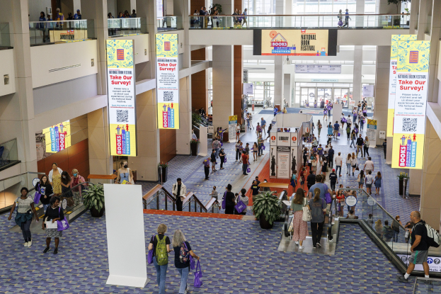 Eine große Menge bewegt sich durch ein großes Gebäude mit Bannern, Topfpflanzen, Rolltreppen, Geländern, Säulen, Texttafeln, Glastüren und anderen Elementen, einige tragen Taschen, was auf die Teilnahme an der Nationalen Buchmesse hinweist.