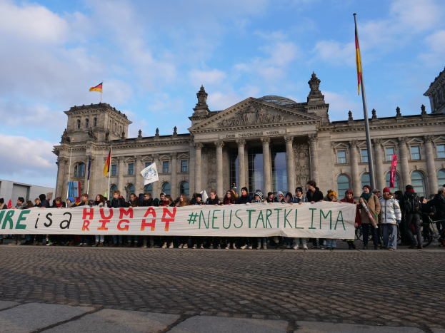 Eine Gruppe von Menschen hält ein Banner mit der Aufschrift "Wir sind ein Menschenrecht" vor dem Reichstaggebäude in Berlin, Deutschland, mit seinen architektonischen Details und umgeben von Fahnenmasten gegen einen bewölkten Himmel.