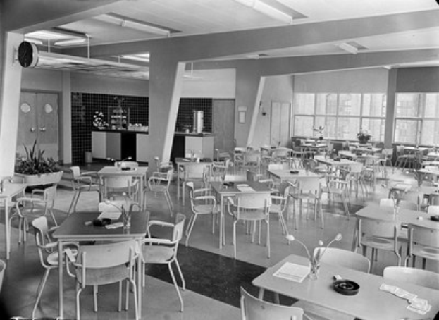 Schwarzes und weißes Foto einer Cafeteria mit Tischen und Stühlen, Tellern, Besteck, Gläsern und Blumenvasen auf den Tischen sowie Türen, Fenstern und Deckenleuchten im Hintergrund.