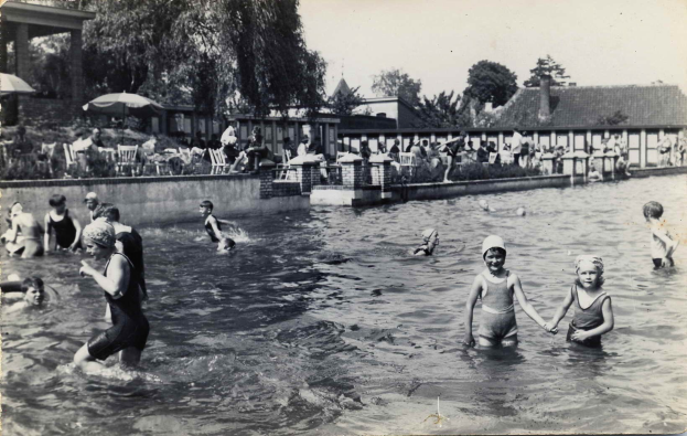 Schwarzes und weißes Foto von Kindern, die in einem Swimmingpool mit Menschen, Sonnenschirmen, Gebäuden und Bäumen im Hintergrund und einem klaren Himmel spielen.