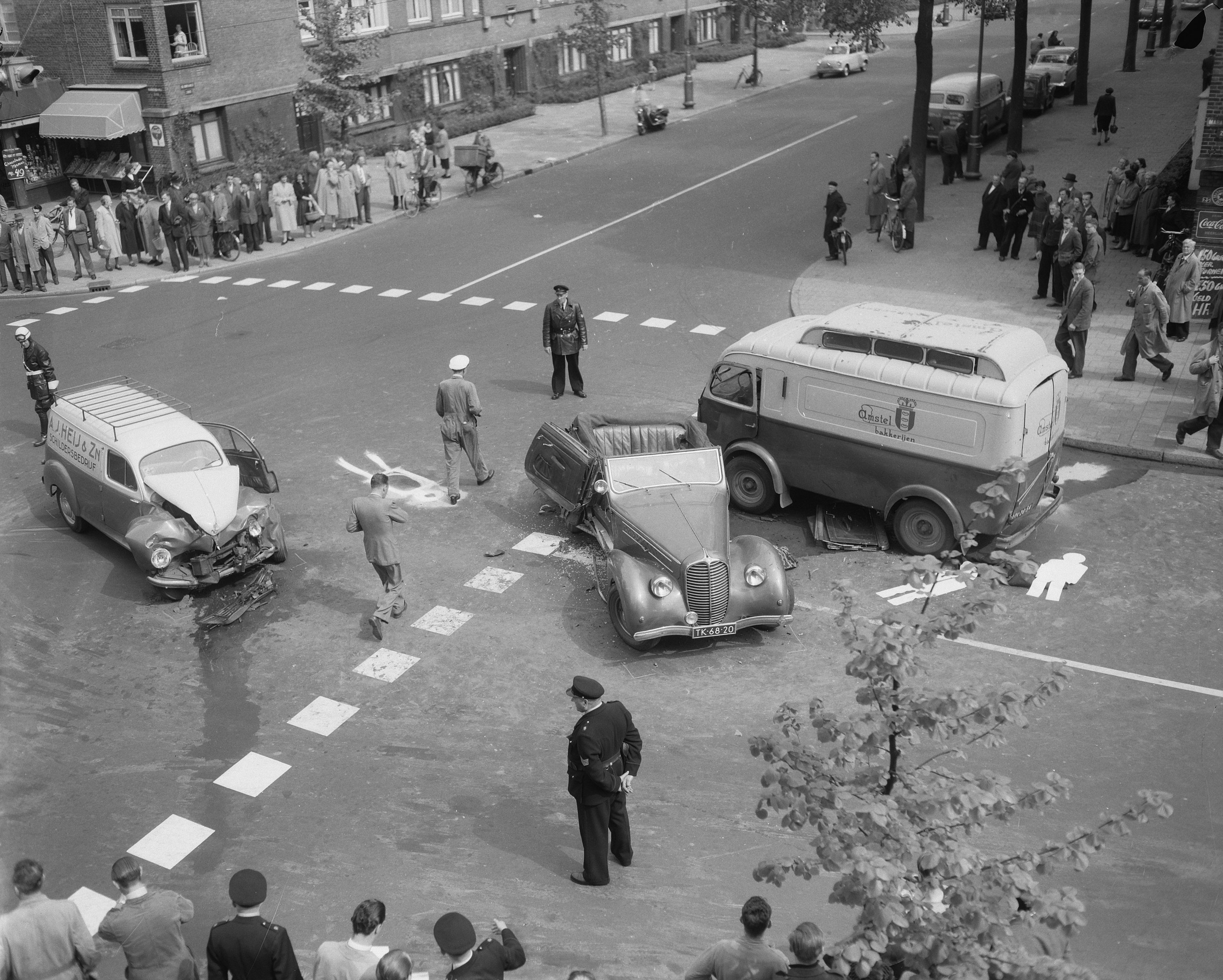 Schwarz-weiß-Szene eines Autounfalls an einer Straßenkreuzung mit mehreren Fahrzeugen, darunter ein Lieferwagen, und einer Gruppe von Menschen in der Nähe, mit Gebäuden, Bäumen, Polen und Texttafeln im Hintergrund.