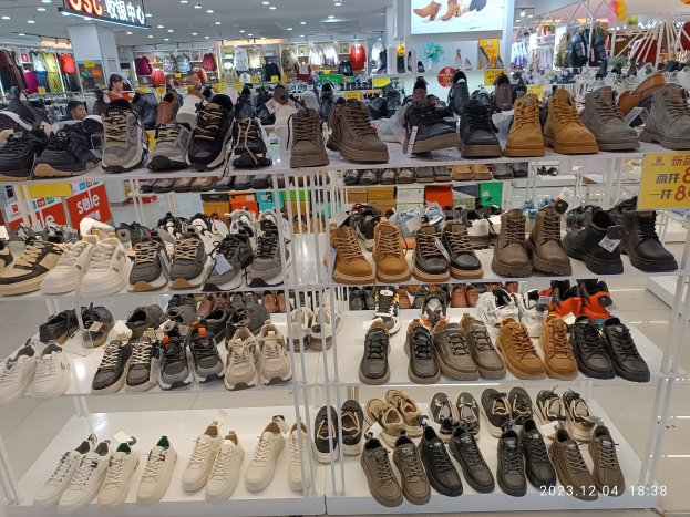 Ein Schuhgeschäfts-Innenraum mit verschiedenen Schuhen auf Ständern, Preisetiketten sichtbar, Menschen im Hintergrund, Kleiderständer, Deckenlampen, ein Bildschirm und Texttafeln.