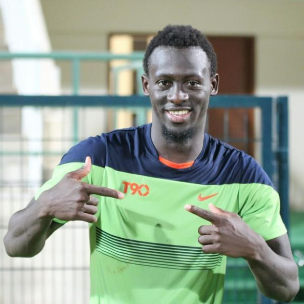 Ein Mann in einem grünen und blauen Hemd, der in die Kamera lächelt und zeigt, mit einem unscharfen Hintergrund, der eine Reling, eine Wand und eine Tür zeigt.