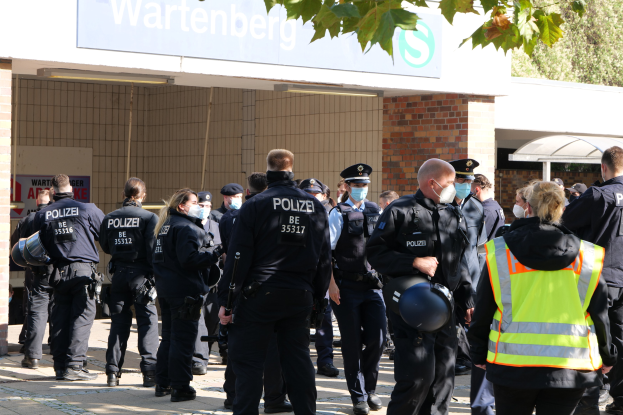 Eine Gruppe von Polizisten steht vor einem Gebäude mit einem Namensschild und Bäumen im Hintergrund, einige tragen Mützen und Masken, andere halten Helme, mit einer Texttafel an der Wand des Gebäudes.