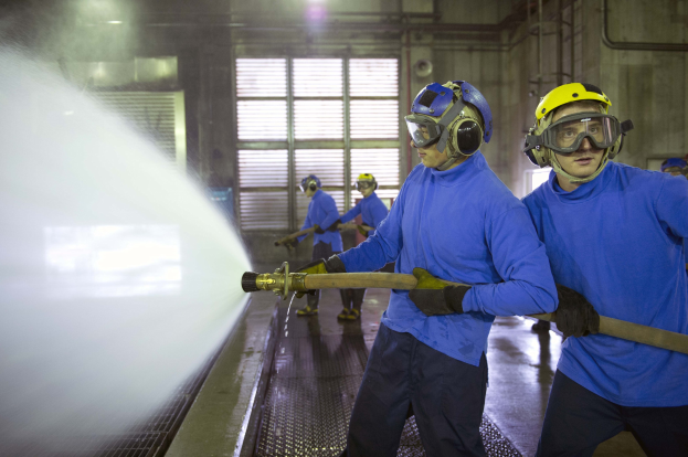 Gruppe von Männern in blauen Hemden und gelben Helmen bei der Arbeit an einer Maschine in einer Fabrikumgebung mit sichtbaren Rohren, Fenstern und Lampen.