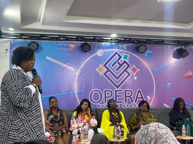 Frau steht vor sitzenden Frauen, hält ein Mikrofon, mit Flaschen und Gegenständen auf Tischen und einem Banner mit Text im Hintergrund.