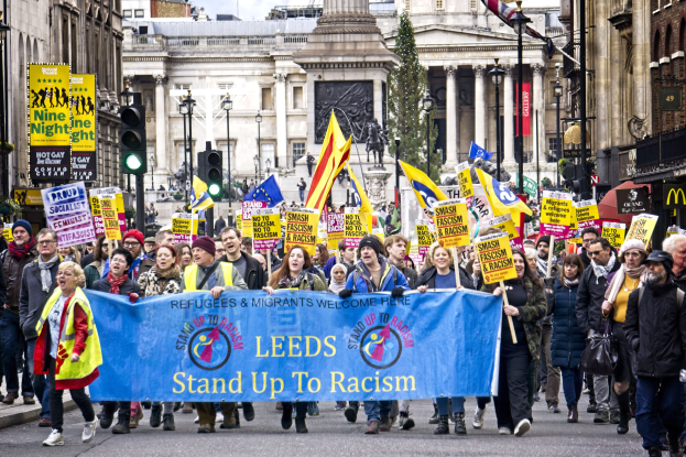 Eine Gruppe von Menschen marschiert mit einer 'Steh Auf gegen Rassismus'-Schleife die Straße entlang, mit Gebäuden, Bäumen, Laternenmästen, Ampeln und einer Statue im Hintergrund bei einem bewölktem Himmel.