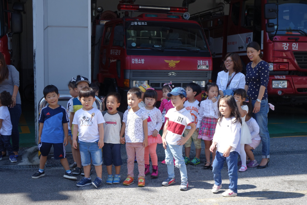 Eine Gruppe von Kindern steht vor einem Feuerwehrauto auf einem Feuerwehrhof, einige tragen Mützen, mit weiteren Feuerwehrautos im Hintergrund.
