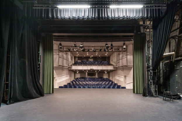 Großer Zuschauerraum mit schwarzen Vorhängen, einer beleuchteten Bühne und Reihen leerer Stühle, mit Gegenständen auf der rechten Seite, die auf eine Veranstaltungslocation hinweisen.