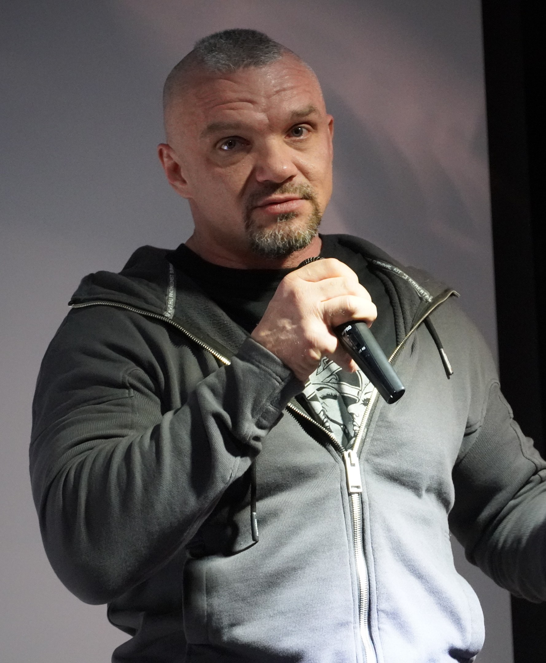 Ein Mann in einer schwarzen Kapuzenjacke und einem schwarzen Anzug mit weißem Hemd hält ein Mikrofon und steht selbstbewusst vor einer Wand, er scheint ein Komiker mit zurückgekämmtem Haar zu sein.