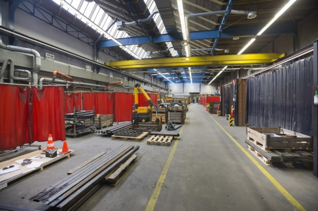 Großer Lagerraum mit Holzpaletten, Metallobjekten, Verkehrskegeln, Maschinenfahrzeug, roten Vorhängen auf der linken Seite, Vorhangwand auf der rechten Seite, Deckenrohre und Lampen.