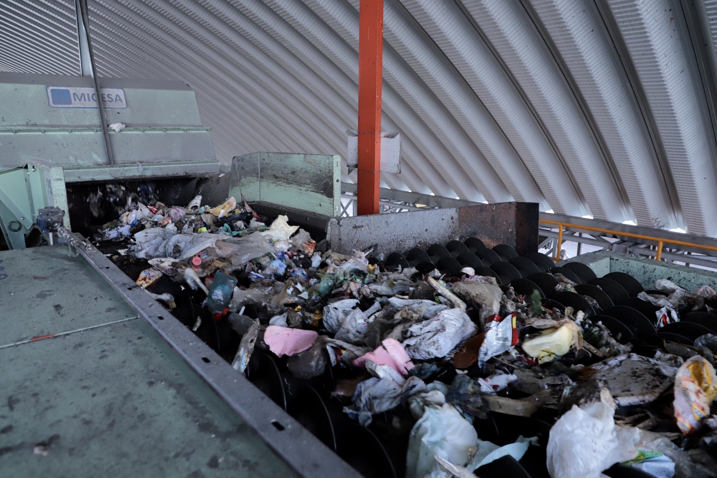 Großer Müllhaufen auf einem Förderband in einer Recyclinganlage mit verstreuter Abfall und einem Metalldach darüber.