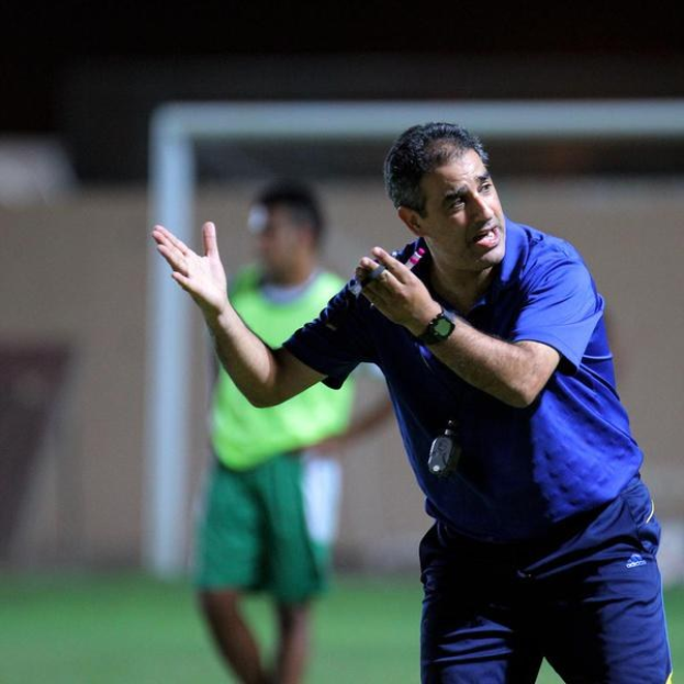 Ein Mann in einem blauen T-Shirt und Hose steht auf einem Fußballfeld und hält ein Mikrofon in der Hand, während er Spielern Zeichen gibt. Im Hintergrund sind eine Person in einem grünen Kleid, ein Tor und eine Wand zu sehen.