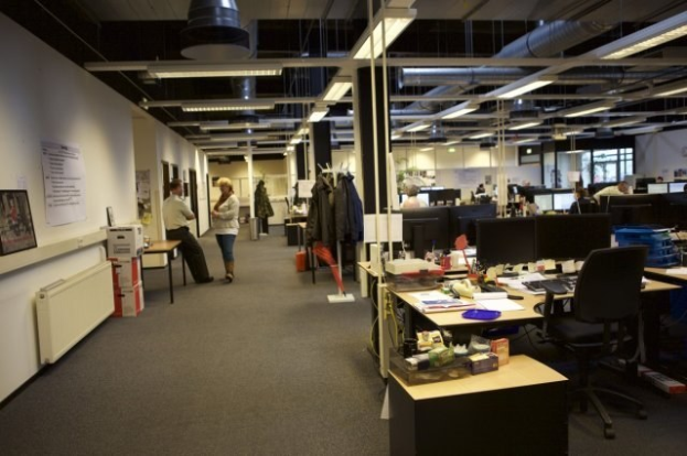 Großraumbüro mit Menschen an Schreibtischen, Computern und Monitoren, umgeben von Papieren, Büchern, Kisten, hängenden Kleidern, wandmontierten Tafeln mit Text und Bildern und Deckenleuchten, mit Glasfenstern im Hintergrund.