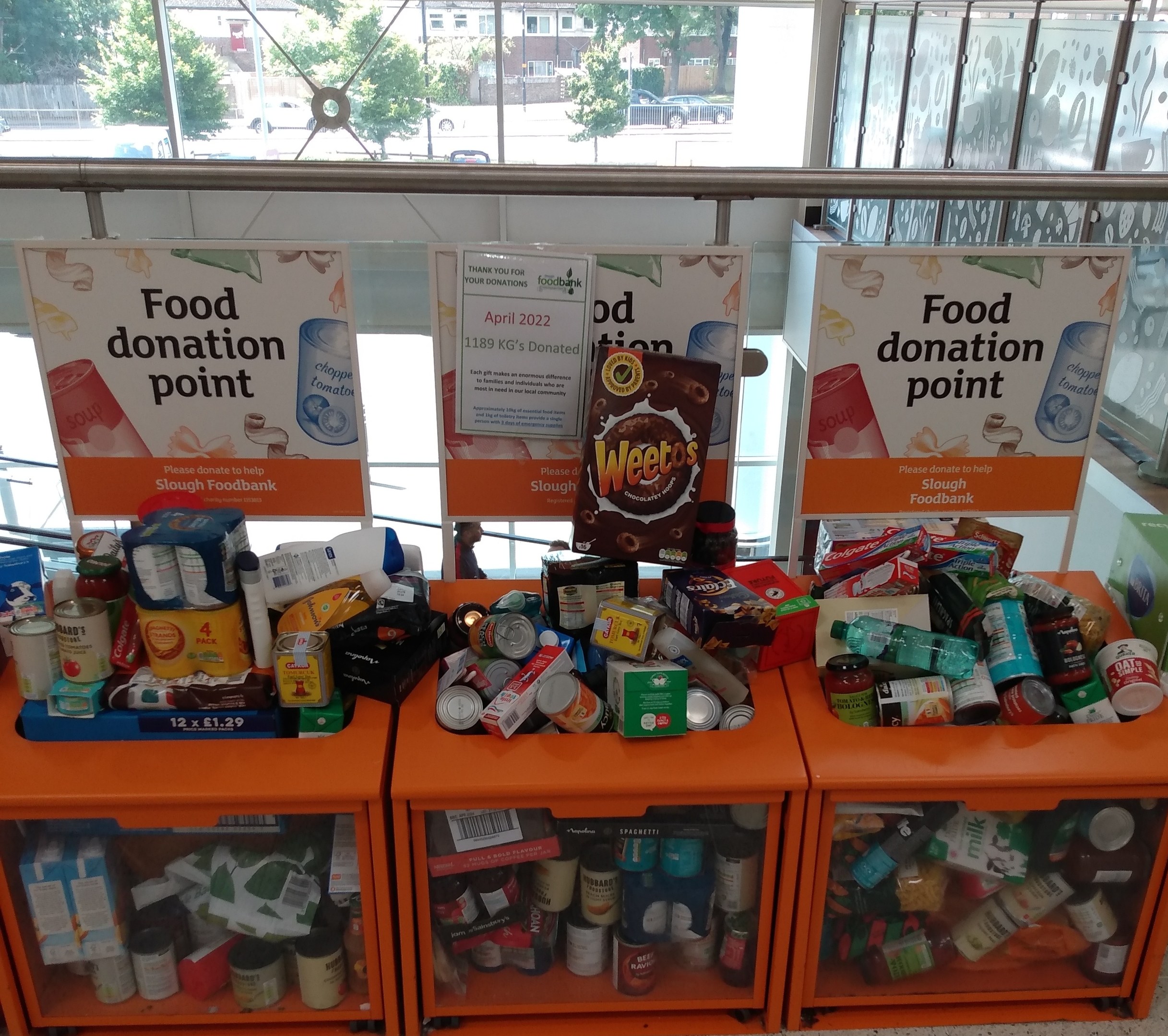 Orange Schränke gefüllt mit Lebensmittelspenden, darunter Flaschen, Dosen und Kartons, mit Informationsschildern und Blick auf Fahrzeuge, Bäume, Gebäude und Himmel durch Glasfenster.