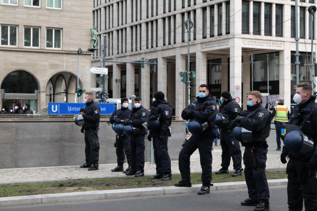 Polizeibeamte in schwarzen Uniformen und Masken stehen vor einem Gebäude mit Glasfenstern und Säulen in Berlin, Deutschland, einige halten Helme, mit Laternen, Verkehrsampeln, Schildern mit Text, einer Statue und anderen Menschen im Hintergrund.