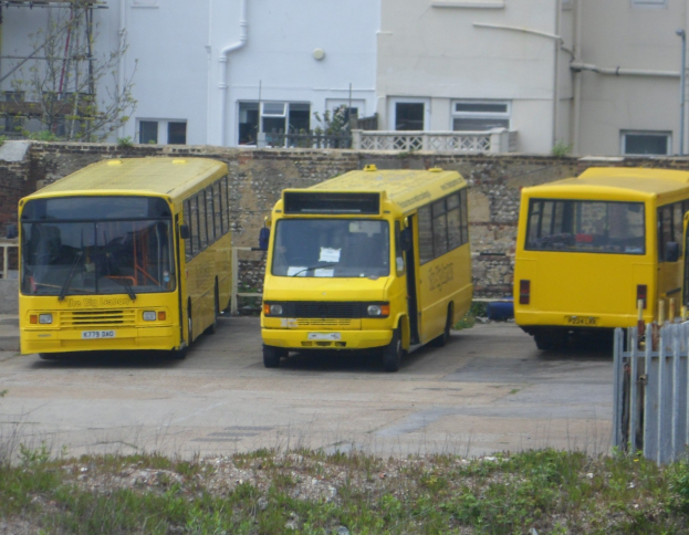 Drei gelbe Busse nebeneinander auf einem eingezäunten Parkplatz mit Gras, einem Gebäude und anderen Strukturen.
