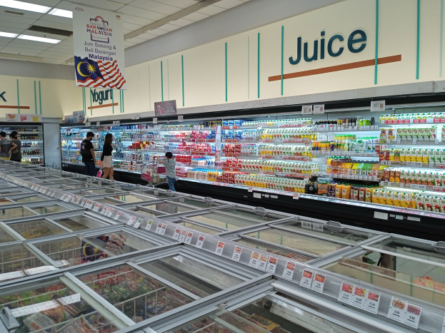 Ein hell erleuchteter Supermarktgang mit einer Vielzahl an Lebensmitteln, darunter Flaschen und frisches Gemüse, sowie einigen Einkäufern und Deckenlampen.