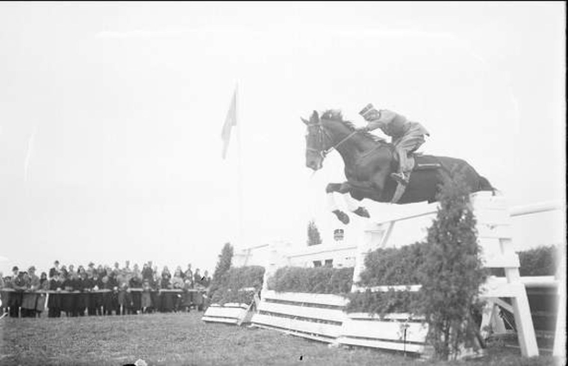 Schwarzes und weißes Foto eines Pferdes und Reiters, die über ein Hindernis springen, bei den 1953 Royal Ascot Horse Trials, mit Zuschauern links, einer Flagge im Hintergrund und Gras unten.