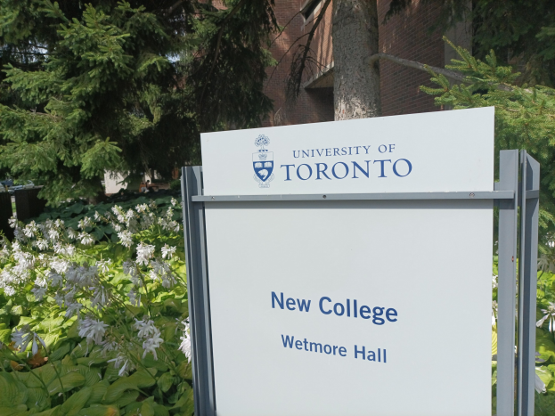 Außenansicht des neuen Colleges der Universität Toronto an Wetmore Hall, mit einem Schild vor dem Gebäude, umgeben von grünem Gras, bunten Blumen, hohen Bäumen und einem klaren blauen Himmel.