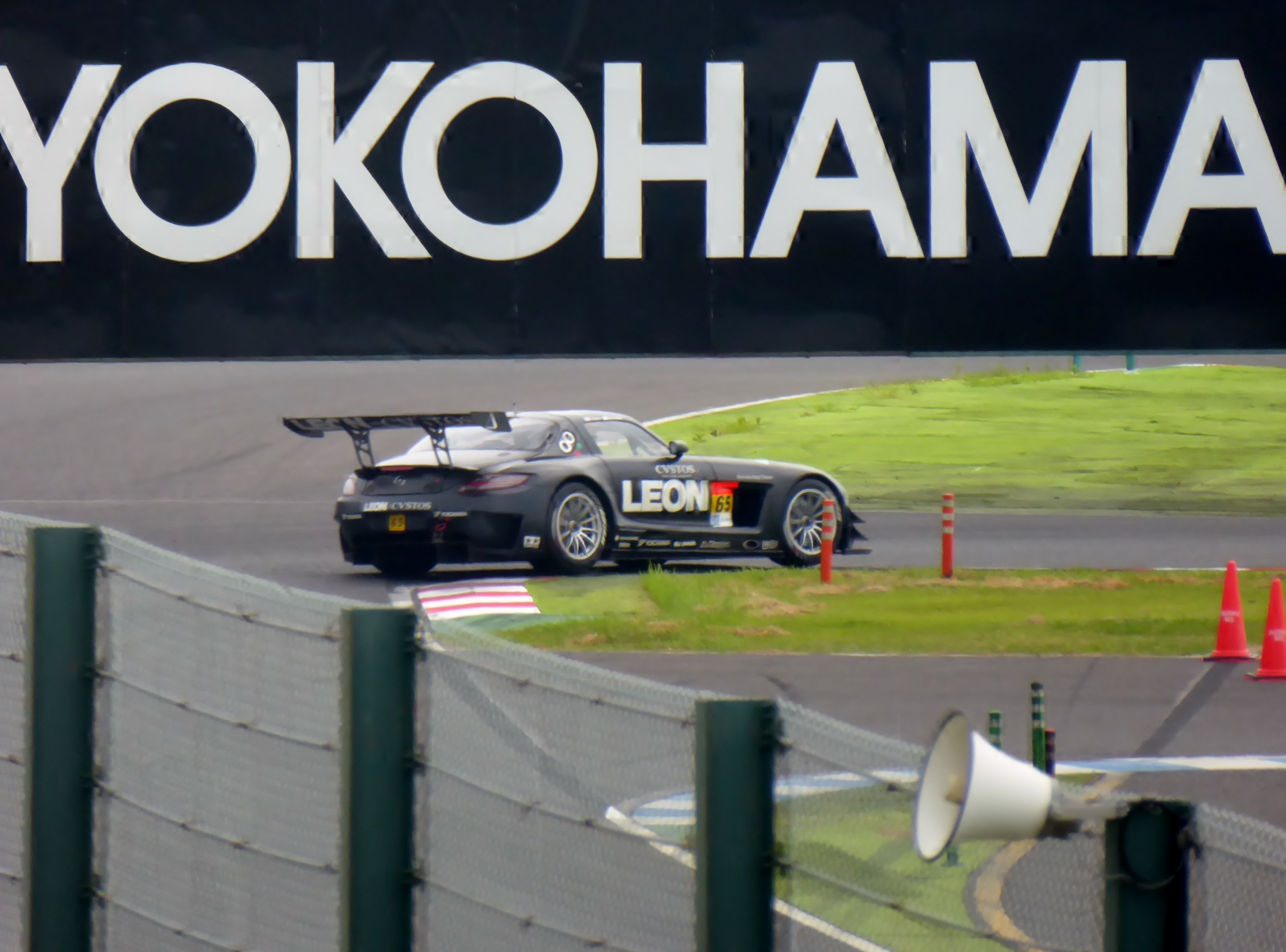 Schwarzer Sportwagen auf einer Rennstrecke mit einem "Yokohama"-Schild im Hintergrund, umgeben von Verkehrskegeln, Gras, einem Zaun und einem Lautsprecher im Vordergrund.