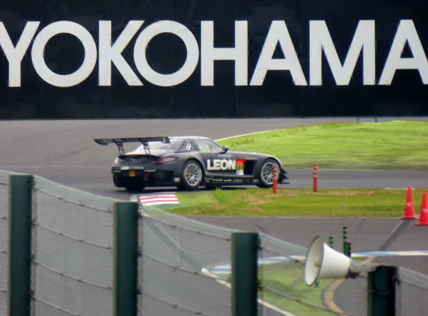 Schwarzer Sportwagen auf einer Rennstrecke mit einem "Yokohama"-Schild im Hintergrund, umgeben von Verkehrskegeln, Gras, einem Zaun und einem Lautsprecher im Vordergrund.