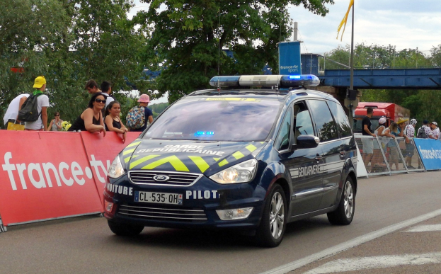 Polizeiauto fahrt auf einer Stra├če neben einer Menge, mit Bannern, Gel├Ąndern, B├Ąumen, einer ├Überquerung, einer Flagge und einem bew├Âlktem Himmel im Hintergrund.