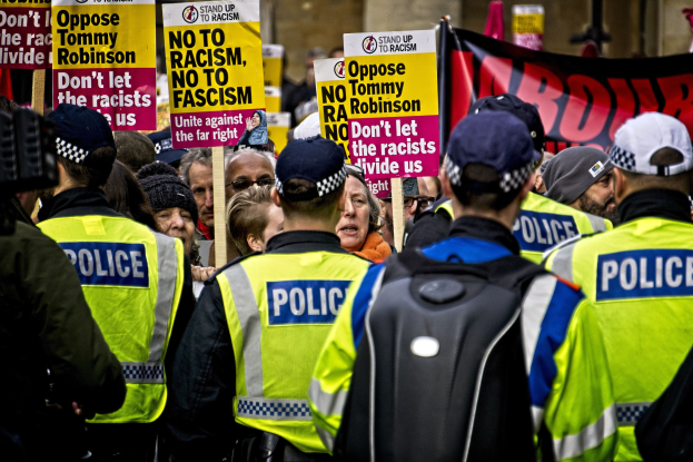 Polizisten vor einer Menge mit Schildern, links ein Filmender und im Hintergrund ein Gebäude.
