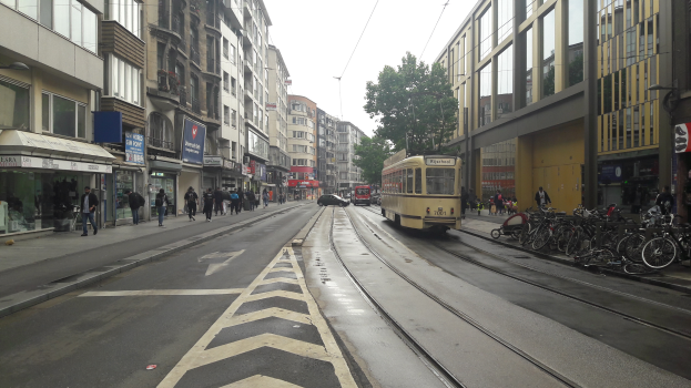 Eine Stadtstraße mit einer gelben Tram, Gebäuden mit Fenstern, Schildern, Menschen auf dem Gehweg, parkenden Fahrrädern, einem Straßenpfahl, Drähten, einem Baum und einem bewölkten Himmel.