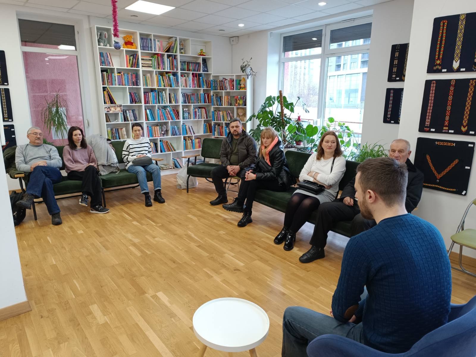 Eine Gruppe von Menschen sitzt um einen Tisch in einem Raum mit Bücherregalen, Pflanzen und einem Fenster, die während eines Workshops zum Thema Gesundheit und Wohlbefinden in Berlin, Deutschland, in ein Gespräch vertieft sind.