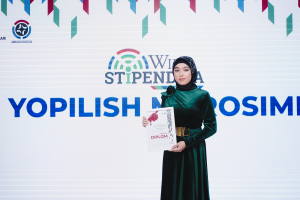 Eine Frau in einem grünen Kleid steht auf einer Bühne und hält ein Zertifikat in den Händen, im Hintergrund ist ein Banner mit der Aufschrift "Yopilish Cosmetics" zu sehen.