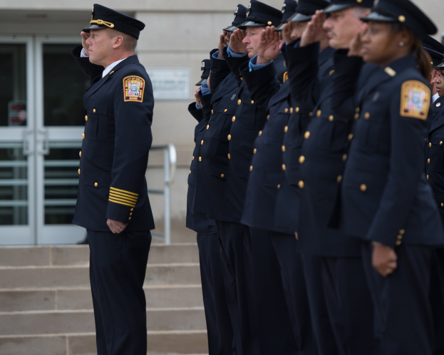 Eine Gruppe von Polizeibeamten in Uniform und Mützen, die in Formation vor einem Gebäude mit Glastüren und Treppen salutieren.