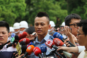 Ein Polizist in Uniform umgeben von Reportern mit Mikrofonen und Aufzeichnungsgeräten, mit Bäumen im Hintergrund.