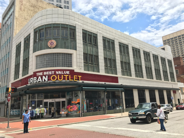 Eine belebte Stadtstraße mit fahrenden Fahrzeugen und Fußgängern auf dem Gehweg, hohe Glasgebäude im Hintergrund und ein sichtbares Schild mit der Aufschrift "The Best Value Urban Outlet" unter einem bewölkten Himmel.