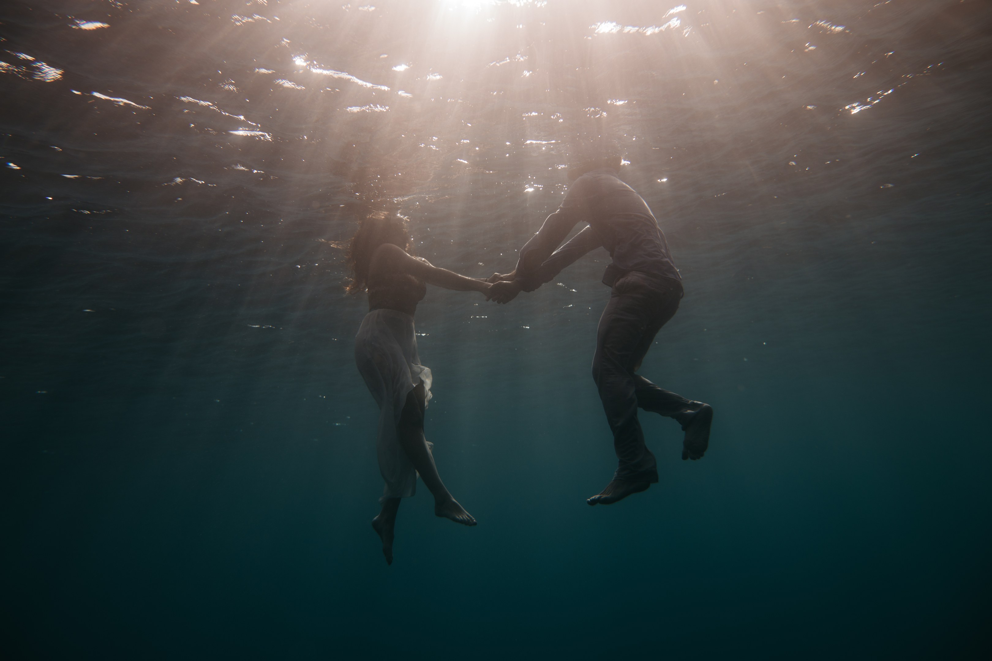 Zwei Personen, die sich unter Wasser in der See an den Händen halten, während Sonnenstrahlen von oben scheinen.