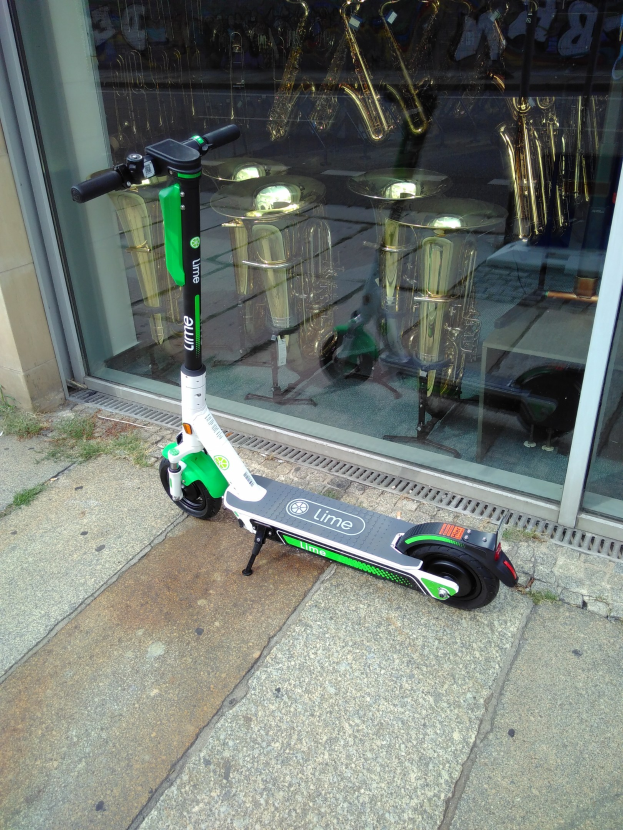 Elektro-Scooter auf einem Gehweg vor einem Geschäft mit einer Glaswand, die Musikinstrumente und andere Gegenstände zeigt.