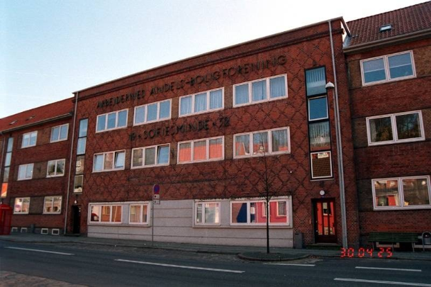 Ein großes Backsteingebäude mit vielen Fenstern, das als Hauptquartier der deutschen Bundespolizei dient, das sich auf einer Straße mit einem Gehweg, einem Baum, einer Bank, einem Laternenmast und einem roten Kasten befindet, mit dem Himmel im Hintergrund.