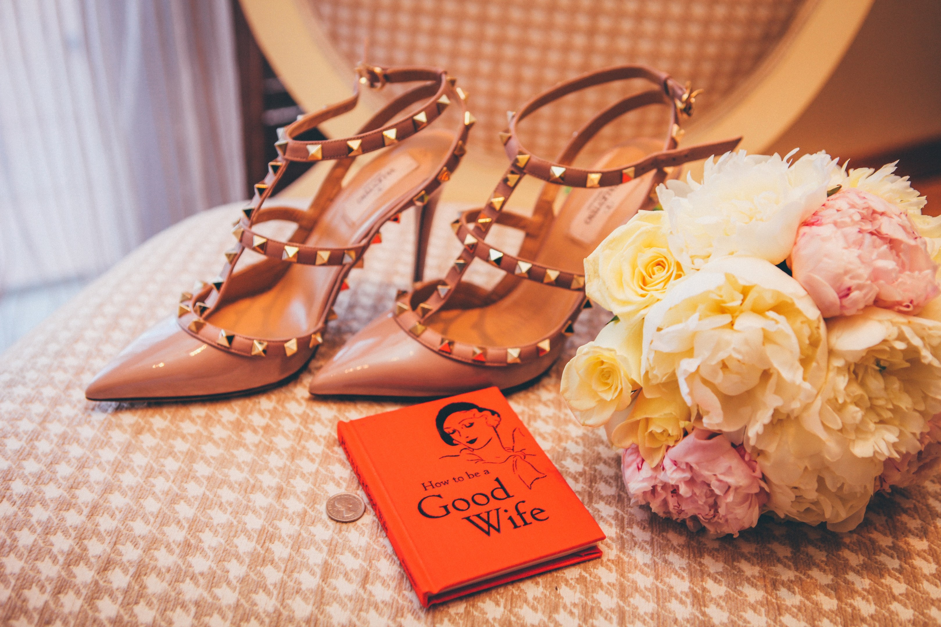 Ein Tisch zeigt ein Buch mit dem Titel "Wie man eine gute Ehefrau ist", einen Blumenstrauß und ein Paar Valentino-Schuhe, vor einem Vorhang als Hintergrund.
