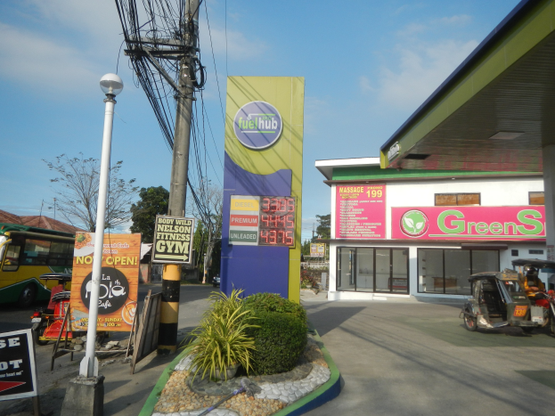 Tankstelle mit einem 'Green's'-Schild, Fahrzeuge auf der Straße, umliegende Pflanzen, Schilder, Pfosten, Lampen, Drähte, Gebäude, Bäume und Wolken am Himmel.