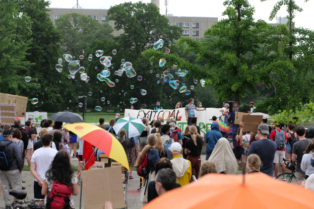 Eine Gruppe von Menschen vor einer größeren Menge mit Schirmen, Taschen und Schildern während einer Demonstration, mit Seifenblasen in der Luft und Gebäuden, Bäumen und einem Turm im Hintergrund unter einem klaren blauen Himmel.