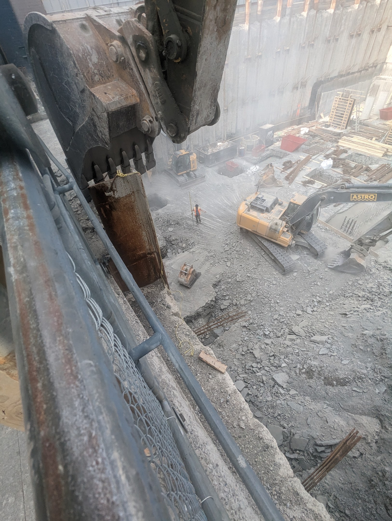 Großer Bagger im Vordergrund auf einer Abrissstelle mit einem Gebäude im Hintergrund, mehrere Fahrzeuge, Personen und ein Metallobjekt links.