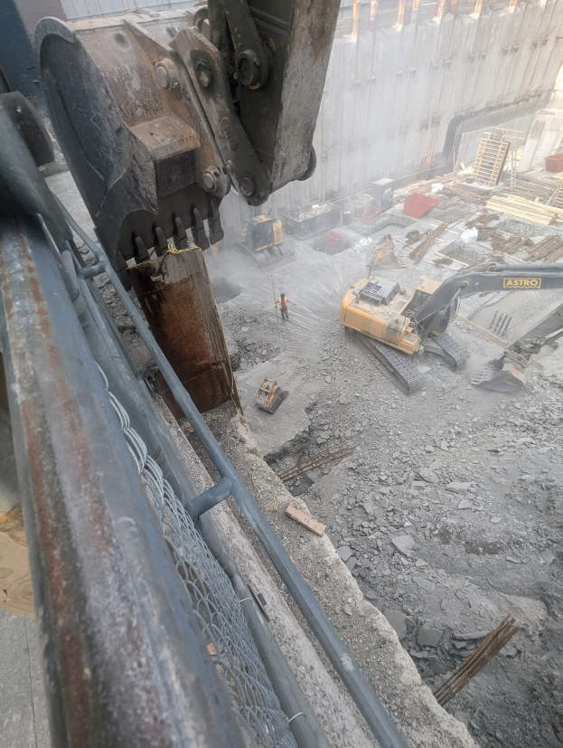 Großer Bagger im Vordergrund auf einer Abrissstelle mit einem Gebäude im Hintergrund, mehrere Fahrzeuge, Personen und ein Metallobjekt links.