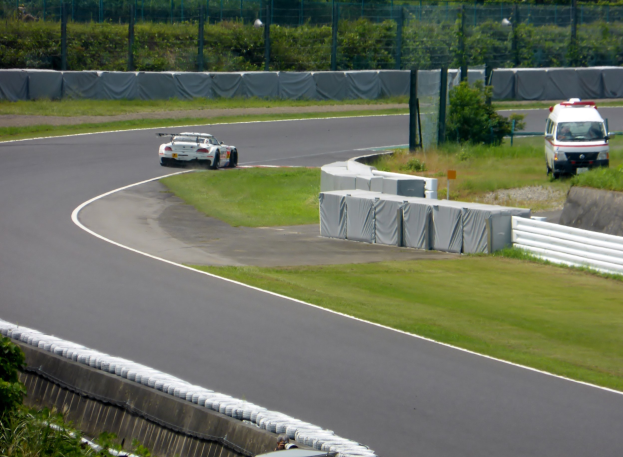 Ein Rennwagen fährt auf einer Rennstrecke mit einem Polizeiwagen im Hintergrund, umgeben von Grünfläche und Absperrungen, mit Text am unteren Bildrand.