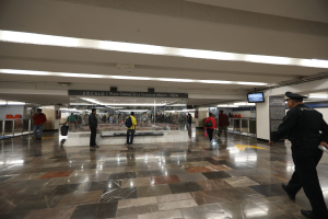 Eine Gruppe von Menschen geht durch eine belebte U-Bahn-Station, mit einem Polizeibeamten im Vordergrund, umgeben von Geländern, Bildschirmen und Schildern.