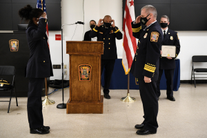 Gruppe uniformierter Personen in Masken auf einem Podium während einer Vereidigung für einen neuen Feuerchief, der ein Zertifikat hält, mit Flaggen, Stühlen, Tischen und einer Wand im Hintergrund.